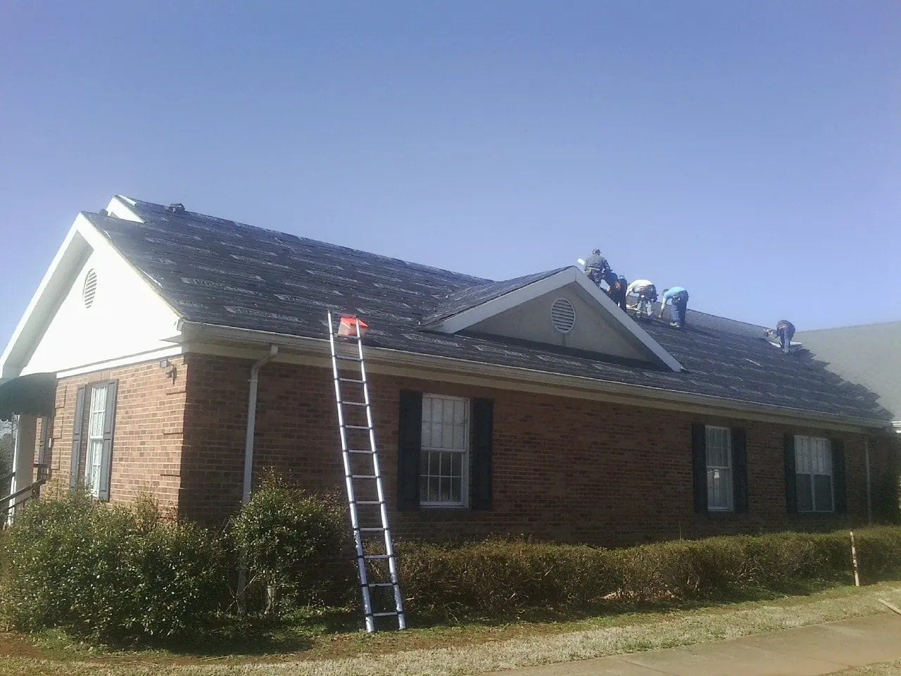 Crew tearing shingles off a brick ranch roof with a ladder set — Emergency Roof Repair work by our crew in Mirror Lake