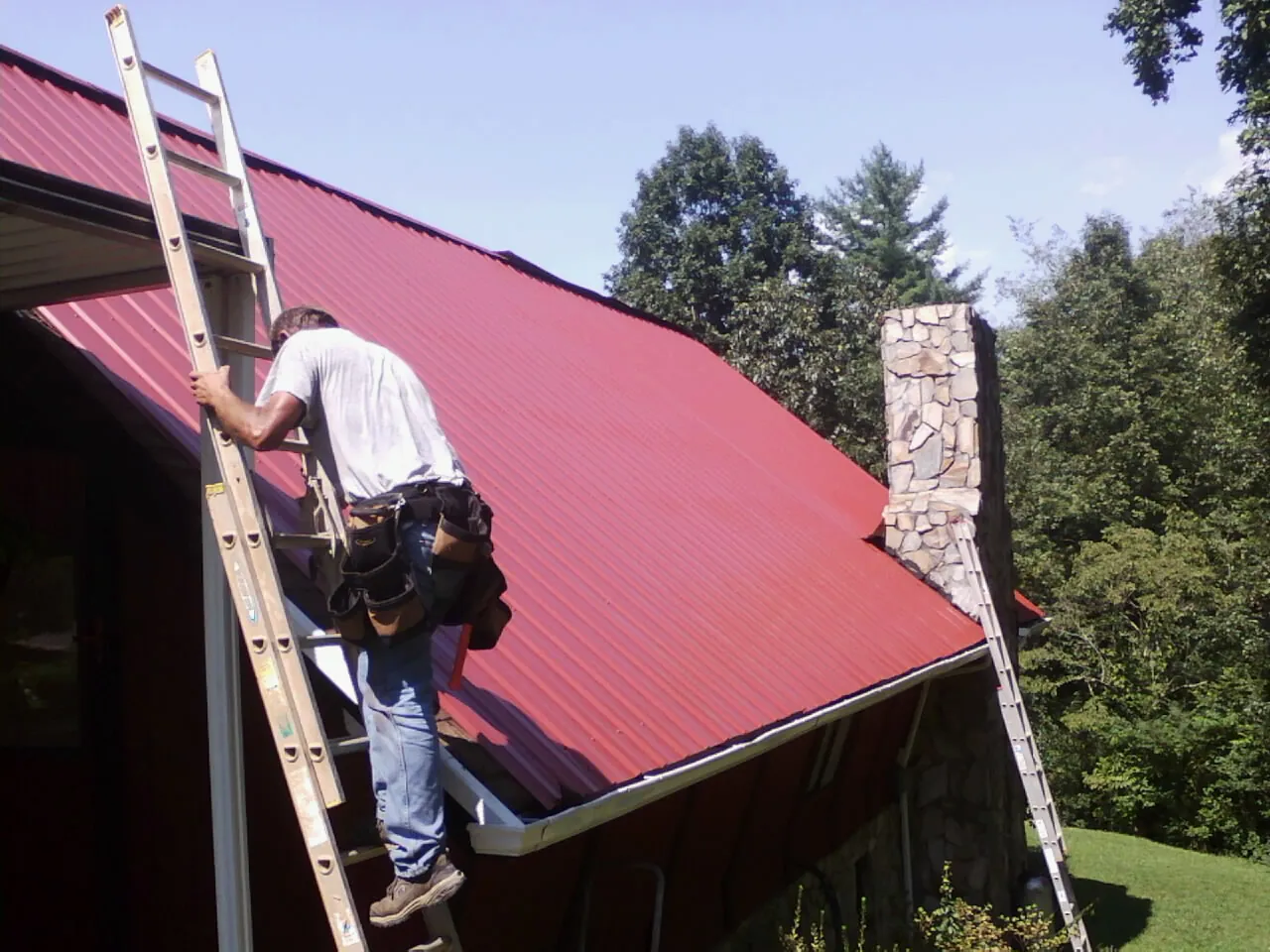 Roofer on a ladder beside a red metal roof and stone chimney — Soffit & Fascia Repair by Roofing Brothers in Mirror Lake